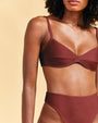 A close-up torso level view of a model wearing a brown bikini top with thin straps and a seam across the middle, including matching high-waisted bottoms. The background is beige.