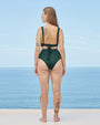 A full body backside view of a blonde model with her hair swept to the side, standing in front of the ocean and the sky, wearing a green two-piece swimsuit with high waisted bottoms and an-open back top with a low bow-tie closure.