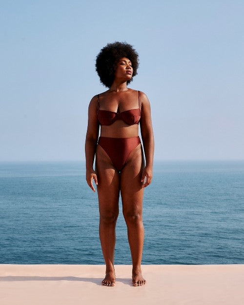 A full-body view of a barefoot model standing on a white surface in front of an ocean and sky, wearing a brown bikini top with thin straps and a structured seam design, including matching high-waisted bottoms.