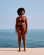 A full-body view of a barefoot model standing on a white surface in front of an ocean and sky, wearing a brown bikini top with thin straps and a structured seam design, including matching high-waisted bottoms.