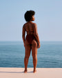 A full-body backside view of a model standing on a white surface in front of an ocean and sky, wearing a brown bikini top with thin straps and matching high-waisted bottoms.
