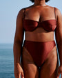 A close-up image of a model standing on a white surface in front of an ocean and sky, wearing a brown bikini top with thin straps and a structured seam design, including matching high-waisted bottoms.