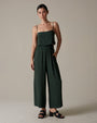 A close-up torso view of a brunette model with her hand on her hip, wearing a dark green spaghetti strapped top with a straight neckline, and matching elastic waistband pants with strapped black open-toed heels. She is standing in front of a grey background.