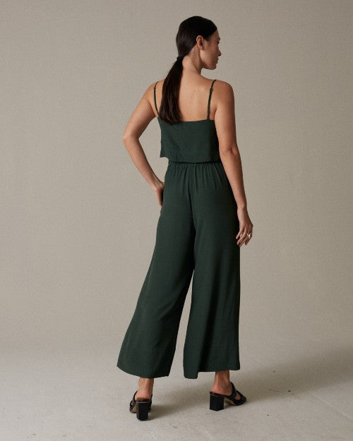 A full body backside view of a brunette model with her hand on her hip, wearing a dark green spaghetti strapped top with matching elastic waistband pants with strapped black open-toed heels. She is standing in front of a grey background.