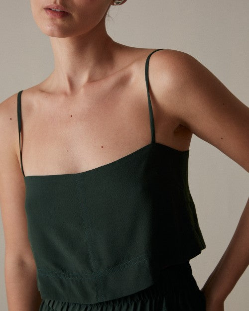 A close-up torso view of a model with her hand on her hip, wearing a dark green spaghetti strapped top with a straight neckline, and matching elastic waistband bottoms. She is standing in front of a grey background.