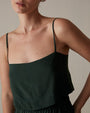 A close-up torso view of a model with her hand on her hip, wearing a dark green spaghetti strapped top with a straight neckline, and matching elastic waistband bottoms. She is standing in front of a grey background.