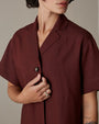 A close-up torso level view of a short-haired model with her hand touching the collar of a maroon short-sleeve button down shirt with brown buttons. The model has tattoos on her forearms and is wearing a silver ring. The background is grey.