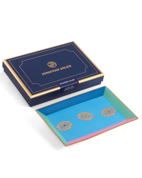 A blue box with an elegant gold trim, a circular logo with the initials "JA" and the text 'JONATHAN ADLER' on the lid. The box is in front of a porcelain multi-color tray with a blue base, gold outlining, and three gold eyes with colorful irises. The background is plain white.