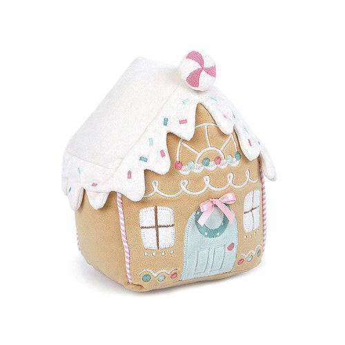 An angled side view of a plush toy gingerbread house with a white icing roof and icing decorations on it, in front of a white background. The roof is topped with a peppermint decoration and colorful sprinkles.