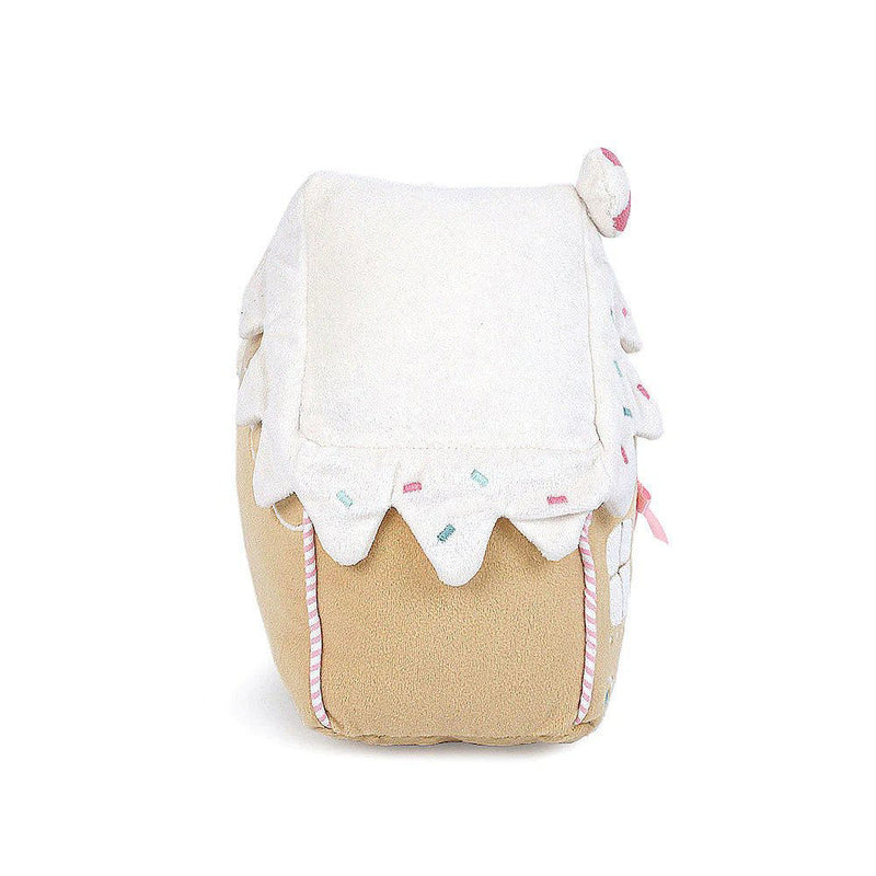 A side view of a plush toy gingerbread house in front of a white background. The house has a white icing roof, decorated with Christmas colored sprinkles and pink candy cane stripes on the sides. 