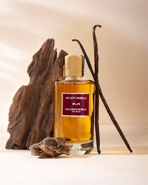 A bottle of Mancera Paris "XPLICIT VANILLA" perfume is displayed with vanilla pods, wood, and resin pieces.