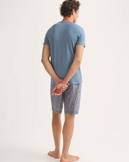 A man in a blue t-shirt and shorts stands turned backwards, appearing casual and comfortable in his attire.