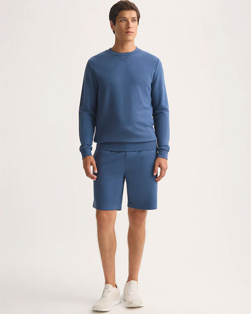 A man dressed in a blue sweatshirt and shorts poses in a relaxed manner.