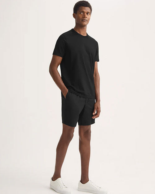A man dressed in a black t-shirt and shorts poses in a casual stance, embodying a laid-back style.