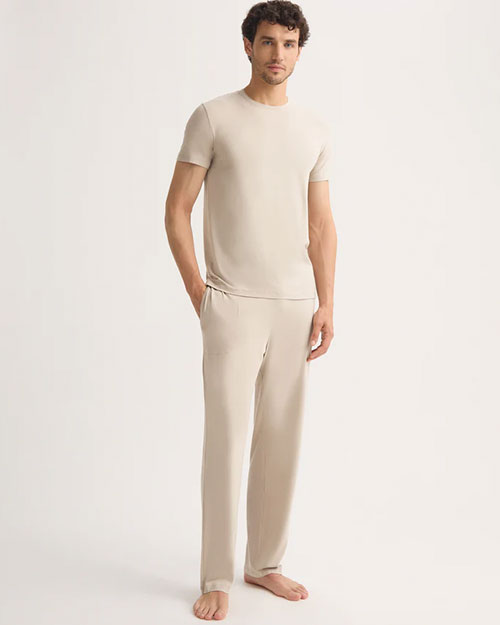 A man wearing a beige t-shirt and pants stands casually, showcasing a relaxed style.