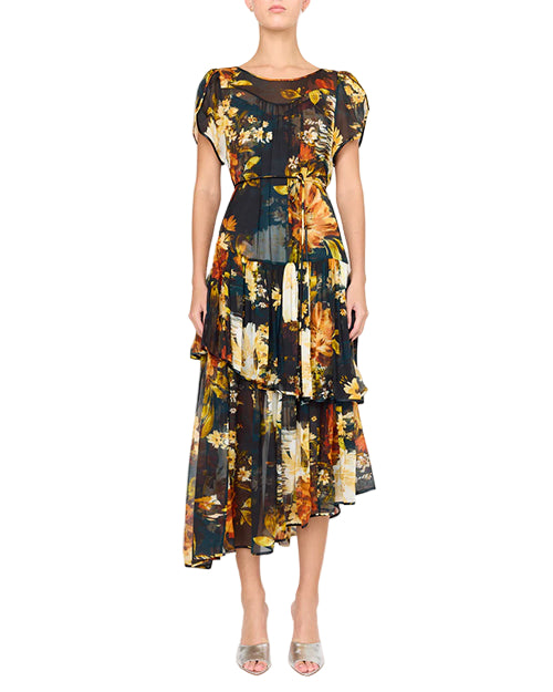 Person wearing a dark floral dress with short sleeves, a round neckline, and an asymmetrical ruffled hemline. The floral pattern features yellow, orange, and green tones. The outfit is paired with open-toed shoes.