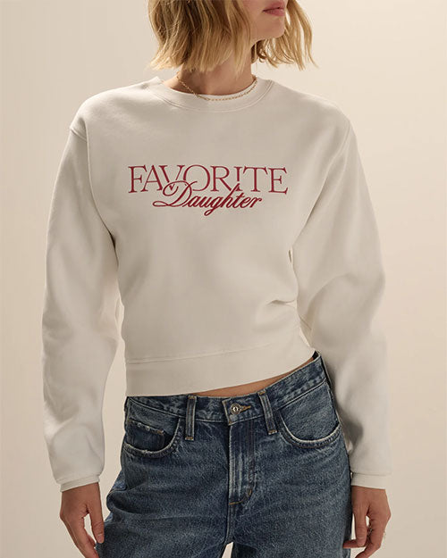 Close-up view of a white cropped sweatshirt with a crew neckline and long sleeves. The sweatshirt features the text “FAVORITE Daughter” printed across the chest in red script and serif lettering and is styled with blue denim jeans. Shown against a light neutral background.