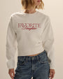 Close-up view of a white cropped sweatshirt with a crew neckline and long sleeves. The sweatshirt features the text “FAVORITE Daughter” printed across the chest in red script and serif lettering and is styled with blue denim jeans. Shown against a light neutral background.