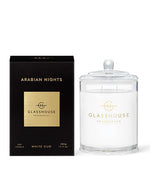 On the left is a black box with gold text that reads ‘ARABIAN NIGHTS GLASSHOUSE FRAGRANCES 380g CANDLE WHITE OUD.’ There is a gold emblem above the text. On the right side of the image, there is a clear glass candle with a gold emblem matching the one on the box, and below it, the words ‘GLASSHOUSE FRAGRANCES’ are visible. The candle has white wax and comes with a glass lid. Both items are set against a plain white background.