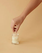 A human hand is shown against a plain, light beige background, reaching towards a small, clear glass candle with white wax inside. The label on the candle reads ‘GLASSHOUSE’ in capital letters.