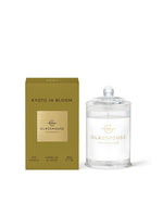 A product from Glasshouse Fragrances named ‘Kyoto in Bloom.’ The image shows a gold-colored packaging box on the left and a candle in a glass container with a lid on the right. The box indicates that it is a soy candle with Camellia & Lotus fragrance, weighing 60g or 2.1 oz.