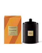 An image featuring a Glasshouse Fragrances candle called ‘In Season Pumpkin Spiced Cake 380g Soy Candle.’ The candle is presented next to its packaging box. The box has a geometric pattern with shades of orange, suggesting an autumn theme. On the front of the box, there is a label with product details in black text. Next to the box is the black glass candle with a gold emblem on the front and a matching black lid on top.