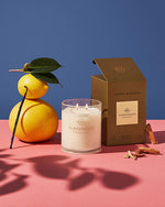 “A still life composition on a dual-tone background with the upper half in blue and the lower half in pink. On the left, there is a yellow citrus fruit, attached to a twig with two green leaves. To its right is an ornamental candle by ‘GLASSHOUSE FRAGRANCES’ with two wicks lit. Adjacent to the candle is its packaging box labeled ‘KYOTO IN BLOOM’ also by ‘GLASSHOUSE FRAGRANCES.’ In front of the box lies pieces of wood scattered around it.