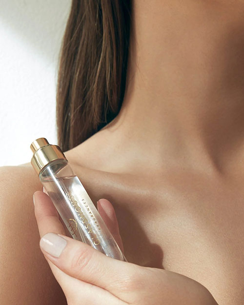 Close-up image of a person’s neck and lower face, holding a small transparent fragrance bottle of liquid with a gold-colored cap. The label on the bottle has 'Glasshouse logo".