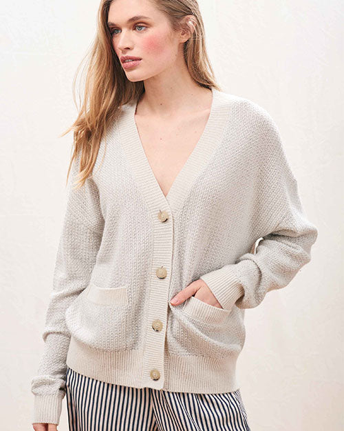 A person wearing a light beige, textured knit cardigan with a deep V-neckline and large front buttons. The cardigan has two front pockets and ribbed cuffs and hem. The person is also wearing high-waisted pants with vertical black and white stripes.
