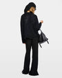 Back view of a woman with a black sweater, black pants, and a black hand bag on a white background.