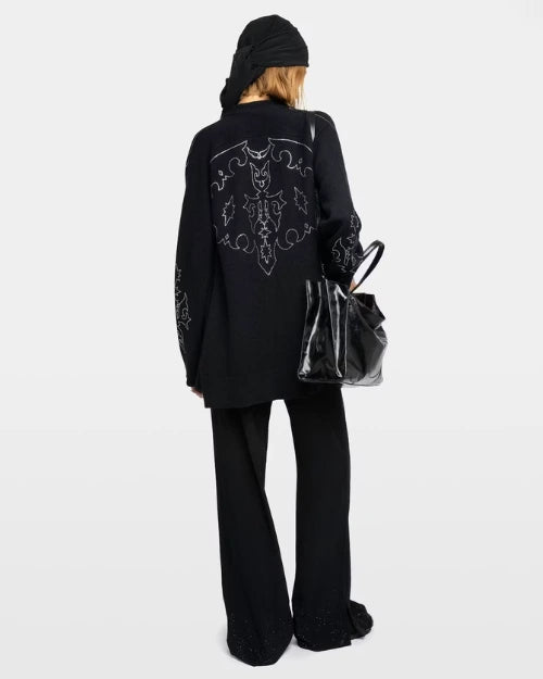 Backside of a woman wearing a black sweater with silver embellishments, black pants, and a black bag against a grey background.
