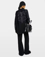 Backside of a woman wearing a black sweater with silver embellishments, black pants, and a black bag against a grey background.