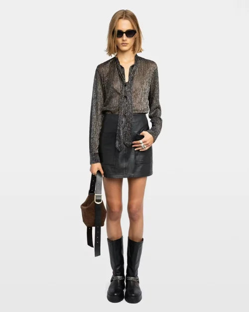 Woman wearing sunglasses with a metallic black shirt, black skirt, black boots, and a brown handbag on a grey background.