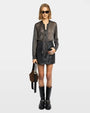 Woman wearing sunglasses with a metallic black shirt, black skirt, black boots, and a brown handbag on a grey background.