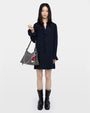 Woman with a navy mini dress and a silver hand bag against a grey background. 