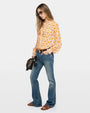 Image of a woman wearing sunglasses, an orange floral blouse, blue jeans, brown sandals and a brown handbag against a white background. 