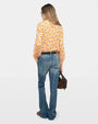 Image of the backside of a woman wearing a pink and orange floral long-sleeved blouse, blue jeans, and a brown handbag against a white background. 