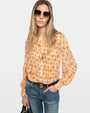 Image of a woman wearing sunglasses with a pink and orange floral long-sleeved blouse and blue jeans against a white background. 