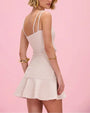 Backside view of a woman with a white mini dress, tied at the waist with rope ties and gold beads on a pink background. 