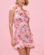 Image of a woman with a pink, floral, ruffled mini dress on a pink background. 