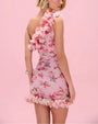 Image of the backside of a woman with a pink, floral, ruffled mini dress with a ruffled hem and neckline on a pink background. 
