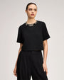 Black short-sleeve top with an embellished jeweled neckline, paired with high-waisted black pleated trousers.