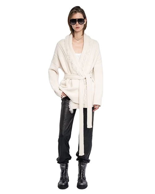Person wearing black sunglasses, a white long-sleeved belted cardigan, black pants, and black boots.