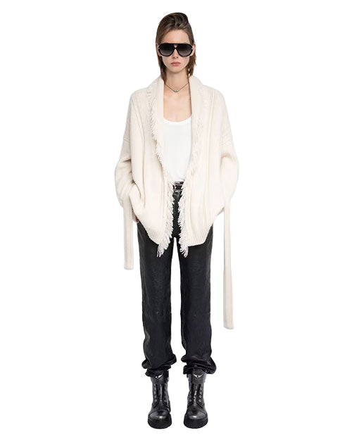 Person wearing black sunglasses, a white fringed cardigan over a white tank top, black pants, and black boots.