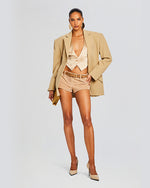 A close-up of nude satin low V-neck top. Top has a vest design with fake pocket stitching and three buttons. Model styles top with tan suede micro shorts, belt, oversized blazer and gold accessories.