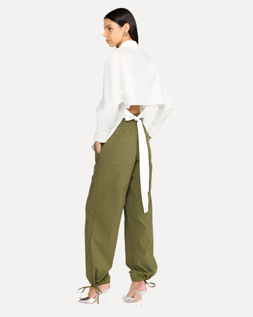 A person is wearing a white long-sleeve top that features an open back design at the lower back. The back/bottom of the shirt is tied together with a delicate bow. The person is wearing green parachute pants with silver heels.