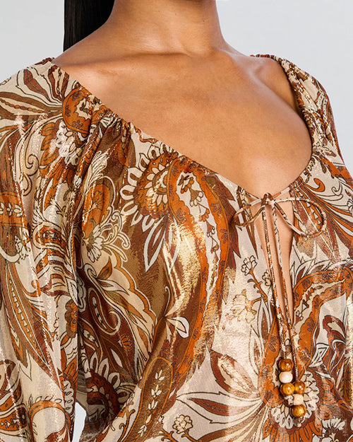 A close-up chest shot of a person wearing a long-sleeved, patterned blouse with earthy tones and a deep neckline. The blouse features intricate floral designs with shades of brown, beige, and orange. 