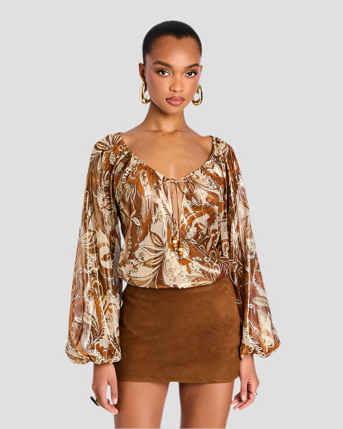 A person is standing against a plain background wearing a long-sleeved, patterned blouse with earthy tones, a deep neckline and a short brown suede skirt. The blouse features intricate floral designs with shades of brown, beige, and orange. 