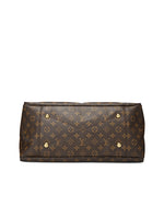 Bottom view of a Louis Vuitton handbag featuring the signature monogram canvas with LV logos and flower patterns in gold tone on a dark brown background. The bag is equipped with four rounded gold-tone metal feet for protection against surface wear, exemplifying the luxury craftsmanship of the brand.