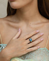 A person wearing a light green lace garment with one hand placed on the chest. The hand features a large oval-shaped blue gemstone ring set in a gold band, and the nails are neatly manicured and painted light pink.